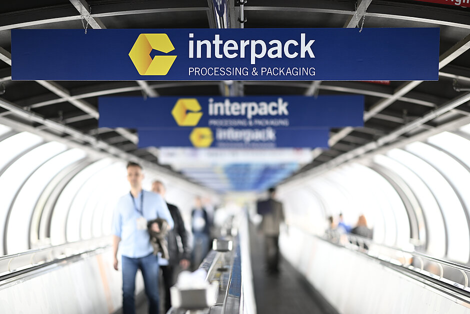 Personas caminando por una cinta transportadora bajo varios carteles azules con el logotipo amarillo de interpack y el texto «interpack PROCESSING & PACKAGING» en una estructura similar a un túnel.